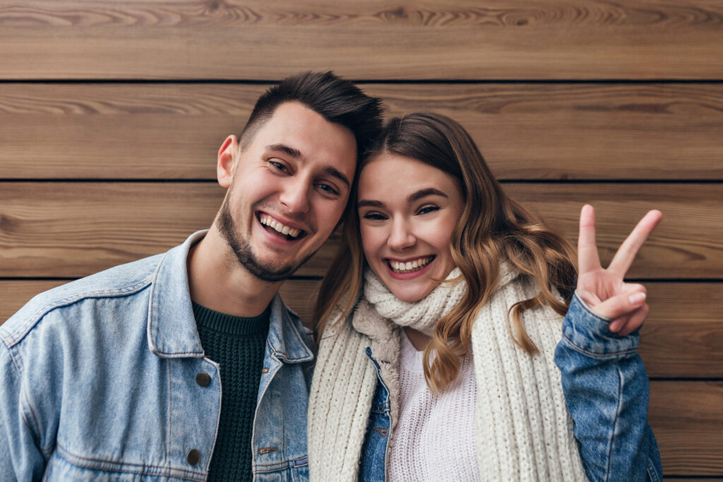 Cheerful european girl in knitted scarf posing with peace sign on wooden background. Indoor shot of laughing married couple having fun together.