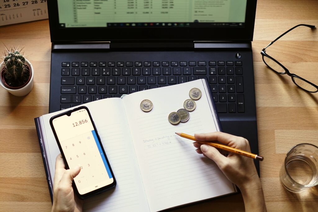 A person calculating loan payments with a smartphone, notebook, and coins in front of a laptop, representing debt repayment planning.