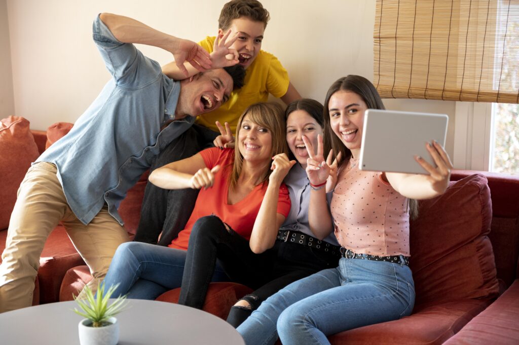 Group of young friends sitting on a couch taking a selfie with a tablet, smiling and having fun indoors in a modern cozy living room with brick wall background, casual happy moment, daytime natural light.