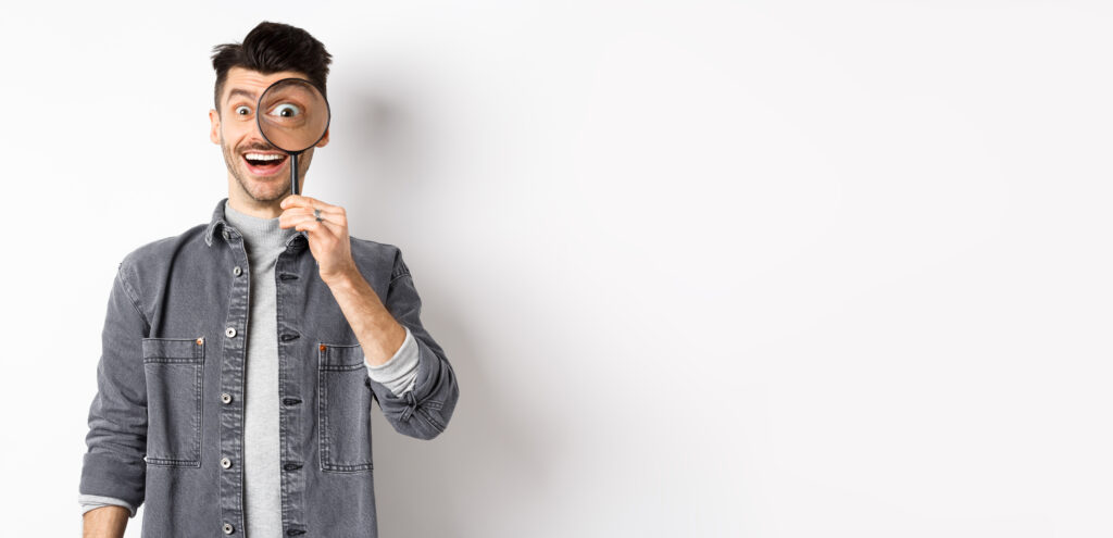 Happy funny guy look through magnifying glass with big eye, checking out interesting deal, searching something, standing on white background.