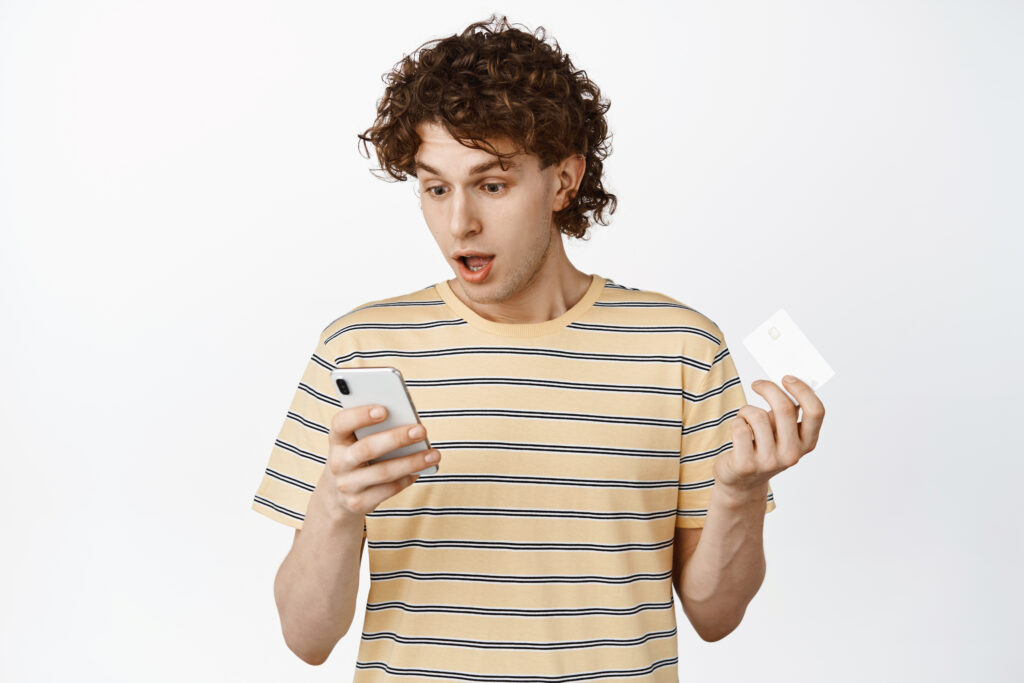 Image of curly guy staring at his phone while placing order, holding credit card, shopping online app, white background.