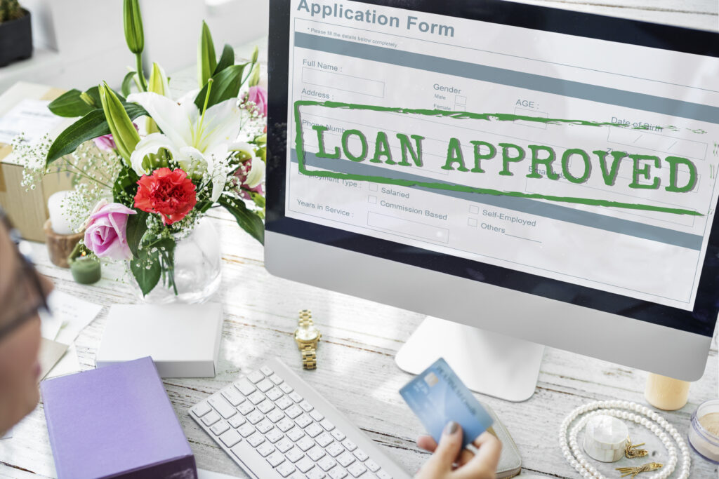 A computer screen displaying a "Loan Approved" message on an online application form, with a person holding a credit card at a desk.