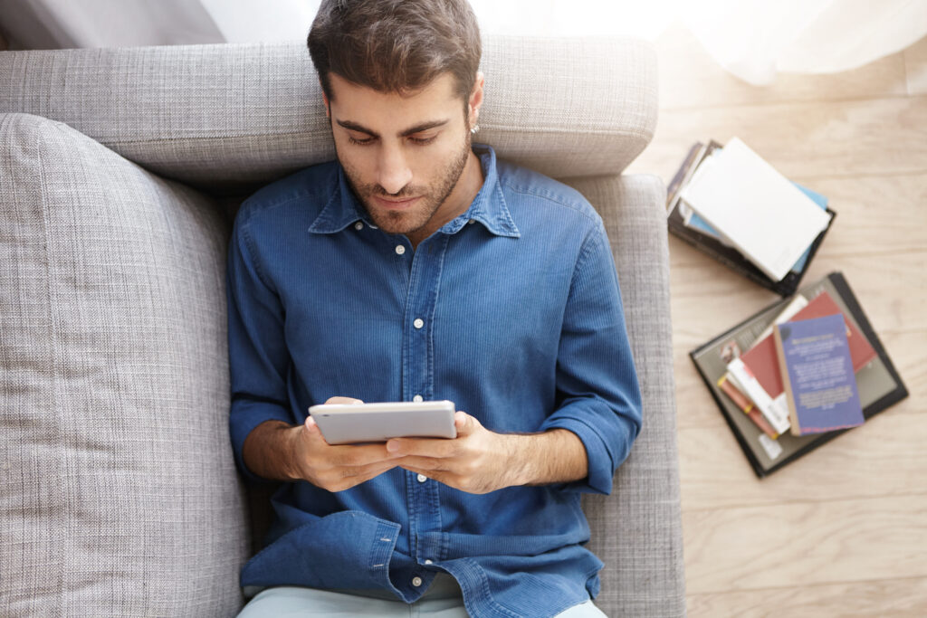Relaxed unshaven male in formal shirt lies on comfortable couch, watches new film on touch pad using free internet connection, plays games online, enjoys conversation with friends in social networks