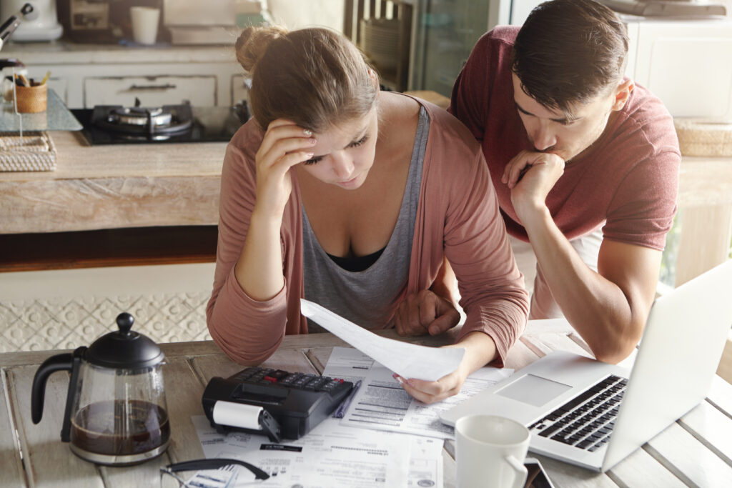 Worried couple reviewing financial documents at home with a laptop, calculator, and papers on the table.