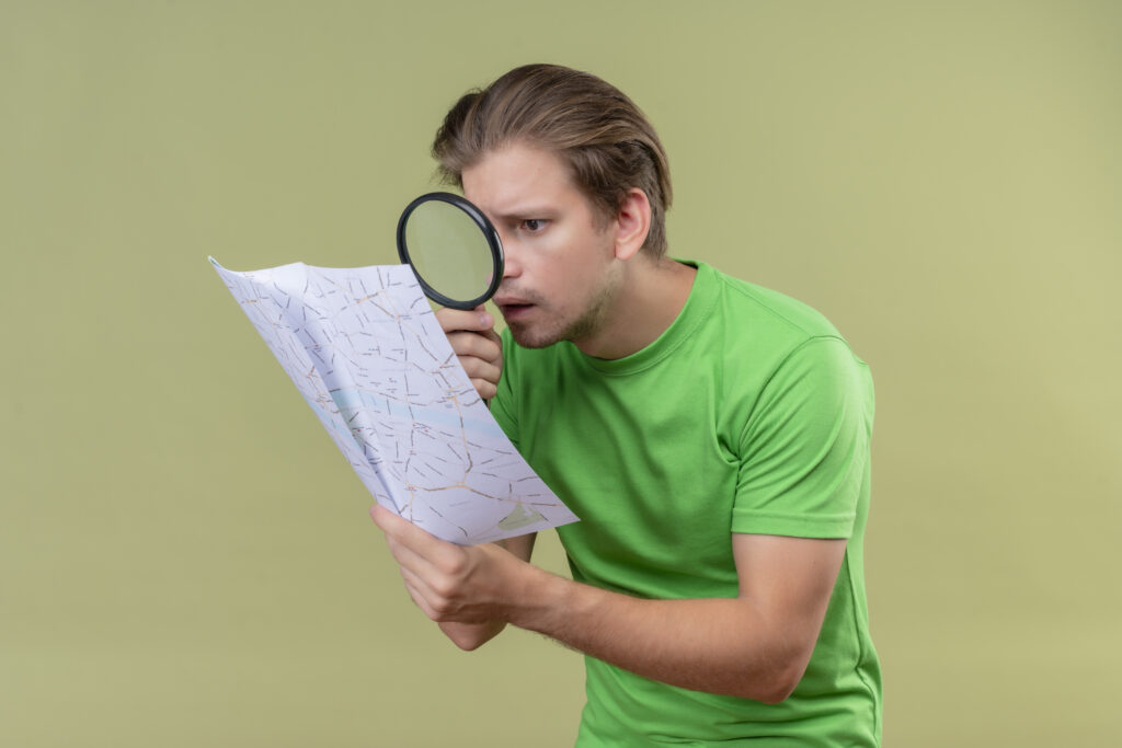 young handsome man wearing green t-shirt looking at map through magnifying glass surprised standing over green background